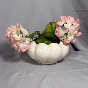 以下が含まれることがあります： ピンクの花と緑の葉が植えられた、波打つ縁のある白い陶器のプランター