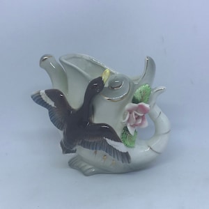 May include: A white ceramic teapot with a duck figurine on top. The duck is brown with gold accents and has a pink rose on its wing. The teapot has a gold rim and a floral design.