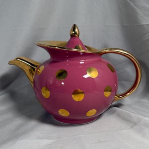 Op de afbeelding: Een roze keramische theepot met gouden details. De theepot heeft gouden stippen, een gouden oor en een tuit. Het deksel is bekroond met een gouden knop. De theepot is ontworpen voor het serveren van thee.