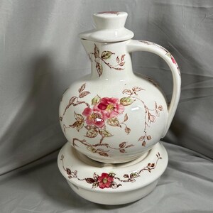 Könnte beinhalten: Weißer Keramik-Krug mit einem Blumendesign mit rosa Rosen und braunen Zweigen. Der Krug hat einen Deckel und steht auf einem passenden Sockel.