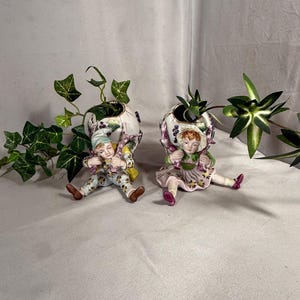 Op de afbeelding: Twee porseleinen beeldjes met zittende figuren, elk met een grote, holle eivormige houder versierd met geschilderde paarse druiven en groene bladeren. De figuren dragen kleurrijke, gedessineerde kleding en hoeden, met klimopbladeren die uit de eivormige houders steken.
