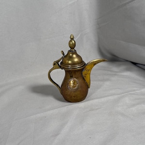 May include: A small, antique brass coffee pot with a curved spout and a handle. The pot has a decorative emblem on the front and a pointed finial on the lid. The pot is made of brass and has a tarnished appearance.