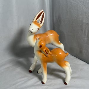 以下が含まれることがあります： 光沢のある仕上げの2頭の鹿の陶器製フィギュアセット。大きい方の鹿は頭を傾けて立ち、小さい方の鹿はその前で上を見上げている。どちらのフィギュアもクリーム色で、耳と蹄に茶色のアクセントが施されている。