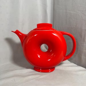 May include: A bright red teapot with a distinctive doughnut-shaped body featuring a central circular opening. The teapot includes a matching lid, a curved handle on the right side, a pointed spout on the left, and a circular base for stability.
