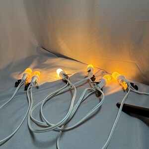Op de afbeelding: Een reeks kleine, warmgekleurde gloeilampen aan witte draden. De lampen zijn gelijkmatig verdeeld en branden, waardoor een zachte gloed ontstaat. De draden zijn verbonden met kleine zwarte clips.