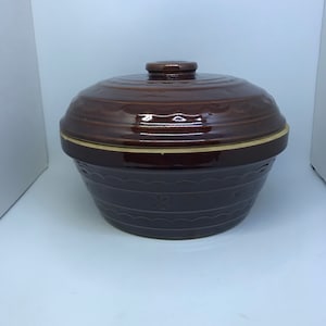 May include: A brown ceramic casserole dish with a lid. The dish has a decorative design around the rim and a snowflake design on the front.