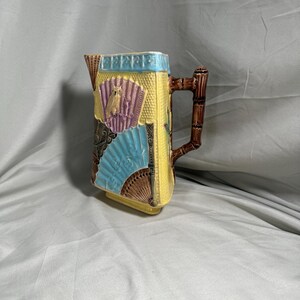 Può includere: Brocca decorativa in ceramica con manico a forma di bambù. La brocca è gialla con disegni di ventagli e insetti in blu, viola e marrone. Il bordo superiore è azzurro. La brocca è probabilmente vintage.