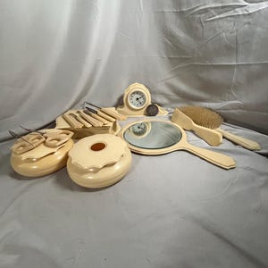 Könnte beinhalten: Ein Vintage-Schminktischset in Elfenbein mit einer kleinen Uhr, einem Handspiegel, einer Bürste, zwei runden Behältern und einem Tablett mit vier kleinen Werkzeugen.