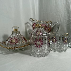 Peut inclure: Un service à thé en verre transparent comprenant un pot à lait, un sucrier avec couvercle et un beurrier avec couvercle. Chaque pièce est ornée d'un motif répétitif de pétales de fleurs magenta et de centres dorés, avec des bords dorés.