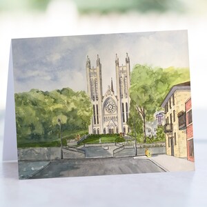 May include: Watercolor painting of a church with two tall towers and a clock face. The church is surrounded by green trees and a street with a building on the right side.