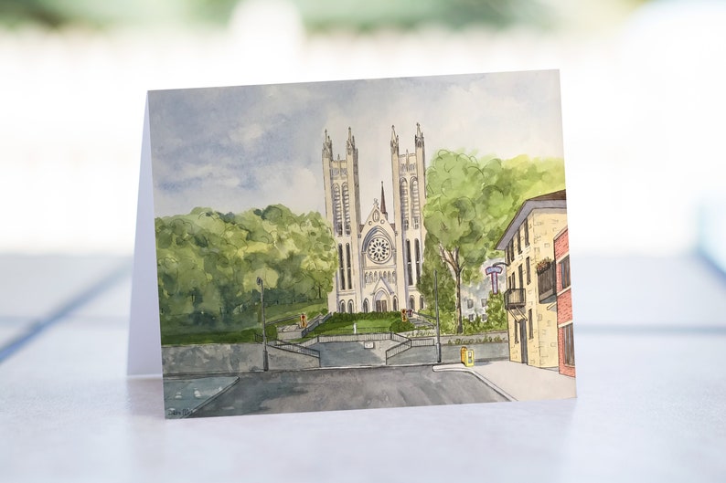 May include: Watercolor painting of a church with two tall towers and a clock face. The church is surrounded by green trees and a street with a building on the right side.