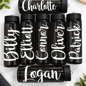 Pu&ograve; includere: Una collezione di sette borracce nere trasparenti con tappi a vite neri. Ogni borraccia presenta un nome bianco stampato in un carattere corsivo. I nomi visualizzati sono Charlotte, Billy, Elliott, Connor, Oliver, Patrick e Logan.