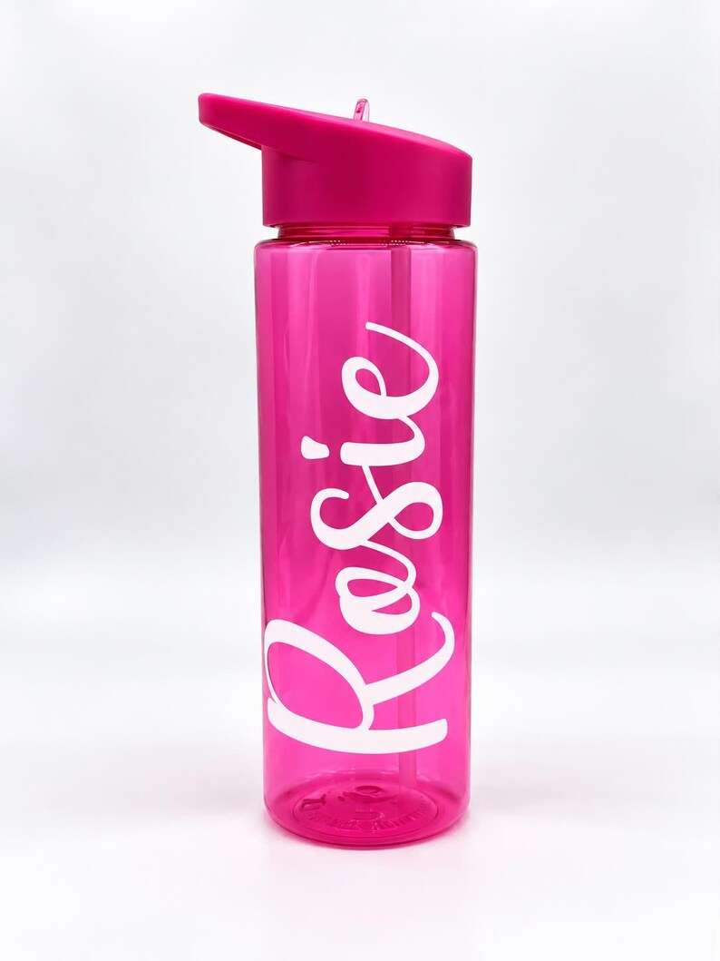 May include: A translucent hot pink water bottle with a matching lid and a flip-top spout. The name Rosie is printed vertically on the side of the bottle in large, white, cursive script lettering. The bottle is cylindrical and made of plastic.