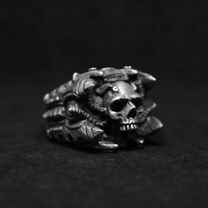 May include: A silver ring with a skull design. The skull is surrounded by intricate details that resemble claws or teeth.