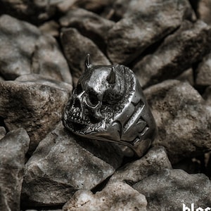 Puede incluir: Un anillo de plata con forma de calavera con cuernos y detalles intrincados. El anillo está sobre un fondo de rocas grises.