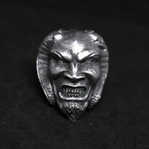 May include: A silver ring with a detailed sculpture of a demon's face. The demon has a fierce expression with sharp teeth and horns.