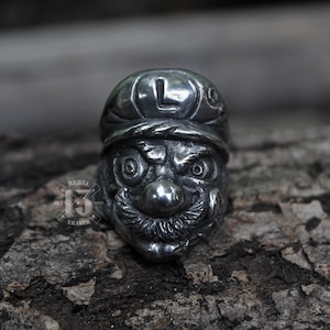 Puede incluir: Un anillo de color plateado que presenta una representación detallada de la cara de un personaje de videojuego. El personaje tiene una nariz grande, cejas pobladas y un bigote. El anillo está colocado sobre un fondo de superficie marrón texturizada.