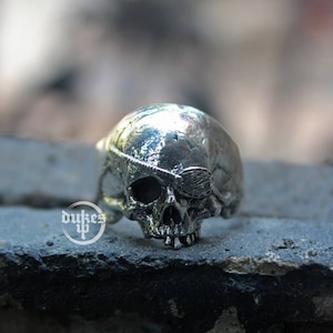 Pode incluir: Um anel de caveira de prata com um tapa-olho. O anel é detalhado com um design de caveira e ossos cruzados.