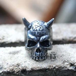Puede incluir: Un anillo de calavera de plata con cuernos y un pentagrama con los números 666 en la frente. El anillo está sobre una superficie gris texturizada. El texto "Rebel 13 Trader" es visible en la superficie.