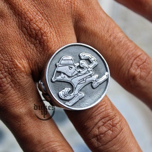 May include: A silver ring with a detailed design of a cartoon leprechaun in a running pose. The ring is worn on a person's finger.