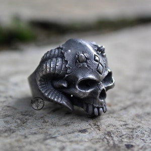 May include: A silver skull ring with horns and a diamond pattern on the forehead.