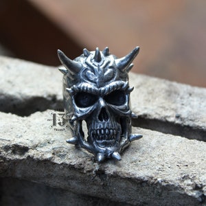 May include: A silver skull ring with horns and spikes. The skull has a detailed design with a menacing expression.