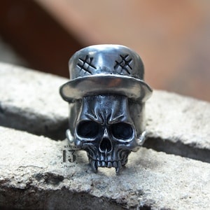 Może przedstawiać: Srebrny pierścionek z czaszką w cylindrze. Cylinder ma na górze wzór w kształcie X. Czaszka ma szczegółową twarz z marszczeniem brwi i ostrymi zębami.