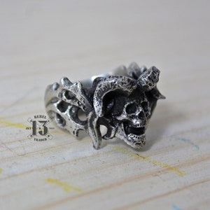 May include: A silver ring with a skull design. The skull has horns and is surrounded by a detailed, intricate design.
