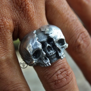 Puede incluir: Un anillo de plata con un diseño de calavera detallado con dos calaveras entrelazadas. Las calaveras están agrietadas y rotas, creando un aspecto único y atrevido.