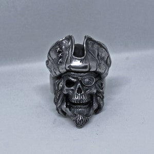 Può includere: Un anello in argento a forma di teschio di pirata con un design dettagliato. Il teschio indossa un cappello da pirata e ha una benda sull'occhio.