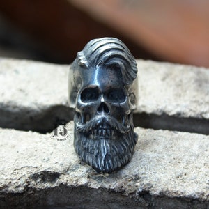 Könnte beinhalten: Ein silberner Ring mit einem detaillierten Totenkopfdesign. Der Totenkopf hat einen Bart und eine Frisur mit Seitenscheitel.