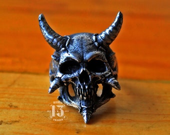 May include: A silver skull ring with horns and detailed features. The ring is on a wooden surface.