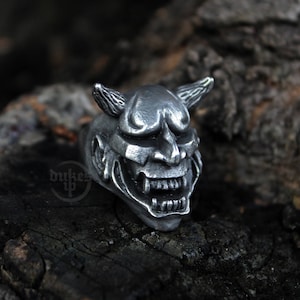 May include: A silver ring featuring a detailed design of a Japanese oni mask. The mask has horns, a wide mouth, and a fierce expression.