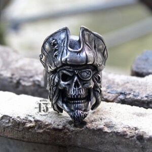 May include: A silver pirate skull ring featuring an eye patch and a tricorn hat. The ring is detailed with a beard and a skull with a menacing grin.