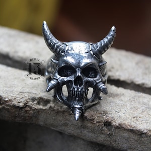 Puede incluir: Un anillo de calavera de plata con cuernos y púas. El anillo está detallado con una cara de calavera y tiene un acabado texturizado.