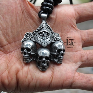 May include: Silver pendant necklace featuring three skulls with an all-seeing eye pyramid design and floral accents. The pendant is attached to a black cord. The pendant also features the text "Rebel 13 Trader 2015".