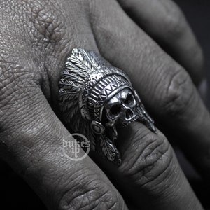 May include: A silver ring featuring a skull wearing a headdress with feathers. The ring is worn on a person's finger.