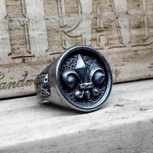 May include: A silver-colored ring with a fleur-de-lis emblem. The ring has a textured surface and a detailed design. The word "Handmade" is engraved on the wooden surface behind the ring. The ring is a piece of jewelry.