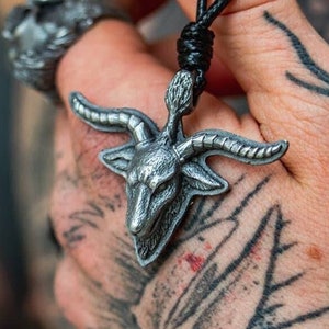 May include: A silver pendant necklace featuring a detailed depiction of a goat's head with horns. The pendant is hanging from a black cord.