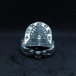 May include: A silver ring with a pyramid design and an eye motif. The ring has detailed carvings, including hieroglyphic-style symbols around the top. The ring's design is reminiscent of ancient Egyptian art. The ring is made of silver-toned metal.