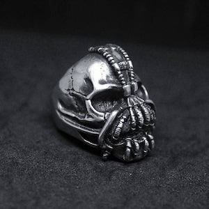 May include: A silver ring featuring a detailed skull design, with a Bane mask and a textured, intricate design.