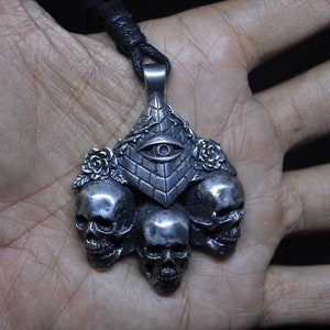 May include: Silver pendant necklace featuring three silver skulls surrounding a silver pyramid with an all-seeing eye design. The pyramid is adorned with vines and roses.