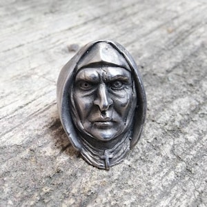 May include: A silver ring with a detailed sculpture of a person's face wearing a hooded robe. The face has a serious expression and the hood is pulled up around the head.