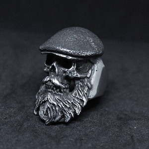 May include: A silver ring featuring a detailed skull with a beard and a flat cap. The skull has a detailed face with a mustache and beard.