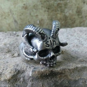 May include: A silver skull ring with horns and the word "sin" engraved on the forehead.
