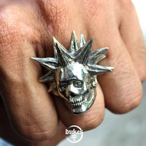 Op de afbeelding: Een zilveren ring met een gedetailleerde schedel met een stekelige mohawk. De ring wordt aan een vinger gedragen.