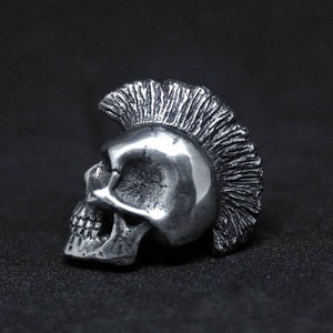 Op de afbeelding: Een zilveren schedelring met een mohawk-stijl haarontwerp. De ring is gedetailleerd met een textuur en is een punkrock-stijl accessoire.