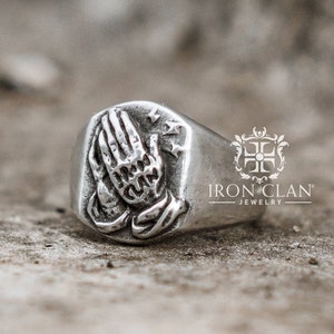 May include: A silver signet ring with a raised design of two hands clasped in prayer. The ring is set on a textured surface.