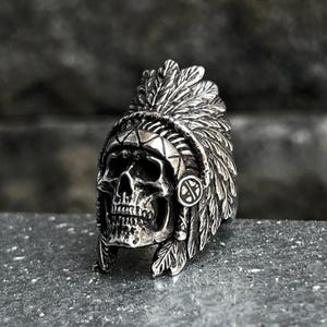 May include: A silver-colored ring featuring a skull wearing a feathered headdress. The skull has detailed facial features and is set against a textured background. The ring's design incorporates Native American cultural elements.