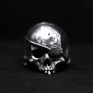 Pode incluir: Um anel de caveira de prata com um tapa-olho. A caveira tem um design detalhado e um acabamento texturizado.